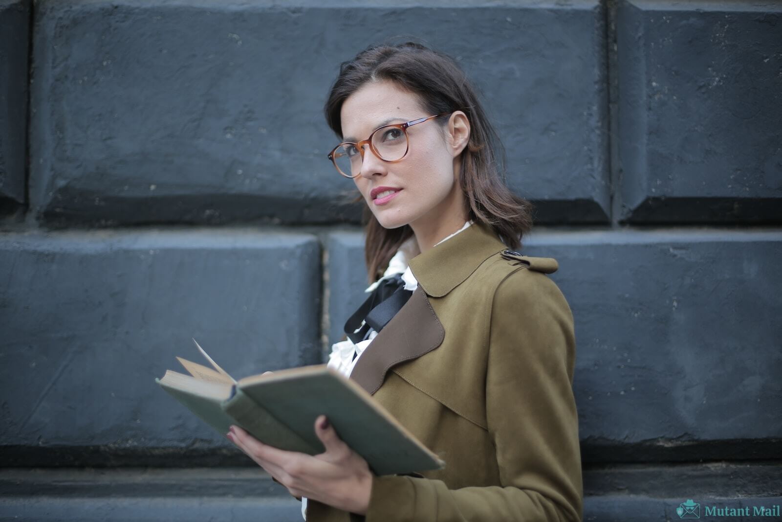 From below confident young female in glasses and elegant vintage clothes looking away and contemplating while reading rook on street near black wall of aged building in city