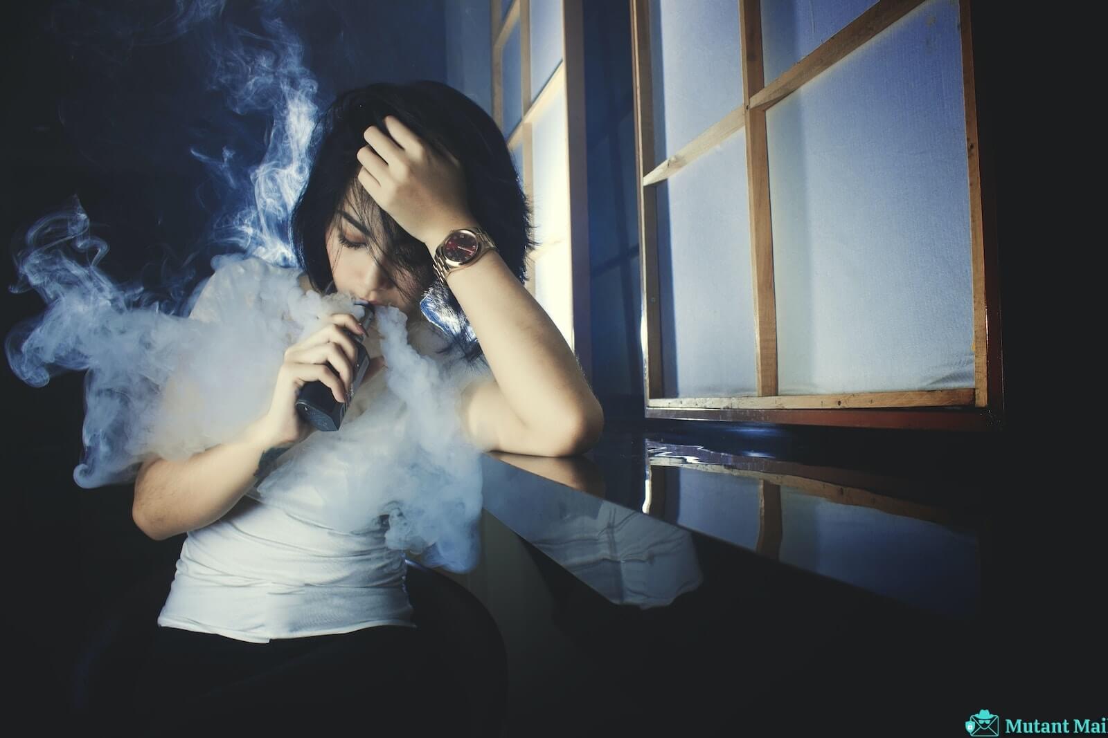 Cool casual brunette in white t shirt smoking electronic cigarette while leaning on hand in dark studio