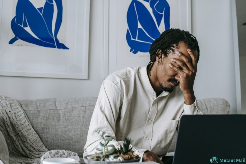Frustrated young bearded African American male freelancer with dreadlocks in casual shirt working on laptop at home and covering eyes with hand after failure